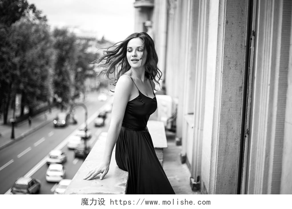 穿着黑色吊带的女人在窗台上眺望黑色白色的年轻女性的肖像在优雅的丝绸黑色礼服看从阳台上的城市, 时尚美容照片